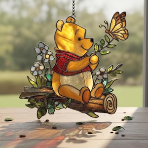 May include: Stained glass Winnie the Pooh suncatcher. The yellow bear is wearing a red vest and sitting on a log with flowers and a butterfly. The suncatcher is hanging from a chain.