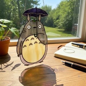 Peut inclure: Suncatcher en vitrail repr&eacute;sentant un personnage de dessin anim&eacute; avec un corps gris, tenant un parapluie violet. Le personnage a de grands yeux et un ventre jaune. Le capteur solaire est suspendu &agrave; une cha&icirc;ne, avec un arri&egrave;re-plan naturel visible.