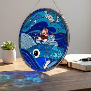 May include: Oval stained glass window depicting a person riding a blue fish through waves and stars. The artwork, framed in dark metal, hangs from a chain. The design features a person with pink hair. The color scheme includes blues, white, and red, creating a vibrant piece.