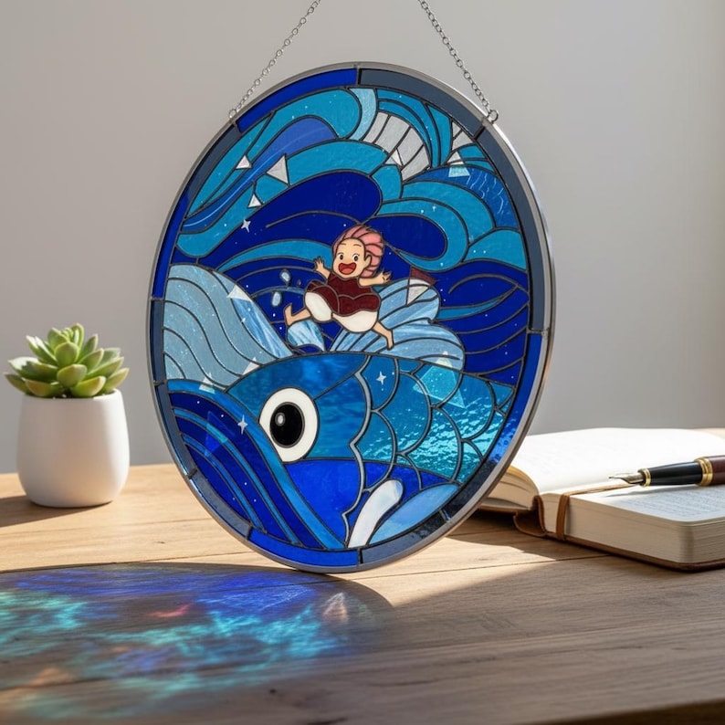 May include: Oval stained glass window depicting a person riding a blue fish through waves and stars. The artwork, framed in dark metal, hangs from a chain. The design features a person with pink hair. The color scheme includes blues, white, and red, creating a vibrant piece.