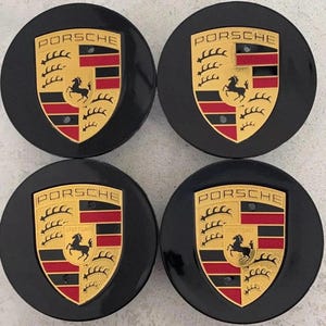 May include: Four black Porsche wheel center caps. Each cap features the Porsche emblem: a gold shield with a black horse, red and black stripes, and the word "PORSCHE" in gold lettering. The word "STUTTGART" is also visible on some caps.