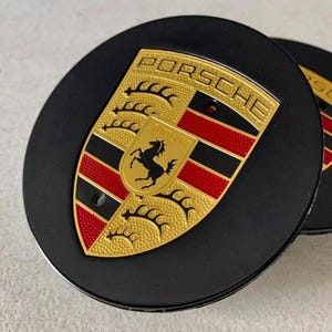 May include: Black wheel center caps with the Porsche emblem. The emblem features a gold shield with the word "PORSCHE" at the top, a black horse, and red and black stripes. The caps are round and have a glossy finish.