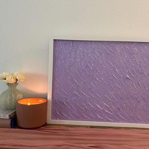 Könnte beinhalten: Ein gerahmtes abstraktes Kunstwerk mit einer strukturierten, lavendelfarbenen Oberfläche. Eine brennende Kerze in einem gerippten, terrakottafarbenen Behälter steht neben einer kleinen Vase mit weißen Blumen und einem Stapel Bücher. Ein rosa Läufer befindet sich im Vordergrund.