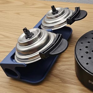 Könnte beinhalten: Nahaufnahme von zwei silbernen und schwarzen Geräteteilen mit einem blauen Halter. Die Teile haben ein kreisförmiges Design mit einer schwarzen Düse und einem schwarzen Griff. Im Hintergrund ist eine schwarze Gerätebasis zu sehen.