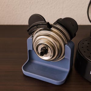 Könnte beinhalten: Nahaufnahme einer Kaffeemaschinenkomponente. Ein silberfarbenes, zylindrisches Teil mit schwarzen Akzenten wird in einem hellblauen Kunststoffhalter gehalten. Ein schwarzer Kaffeemaschinensockel mit dem Markennamen "PHILIPS" ist sichtbar.