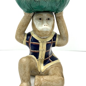 May include: A ceramic monkey figurine holding a green bowl on its head. The monkey is wearing a blue and yellow plaid shirt.