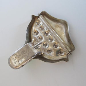 Op de afbeelding: Zilverkleurige metalen citruspers met een handvat en een getextureerd oppervlak. De citruspers heeft een reeks kleine gaatjes om citrusvruchten uit te persen. Het handvat heeft een klein boomontwerp en de tekst "M.A.K." erop gegraveerd.