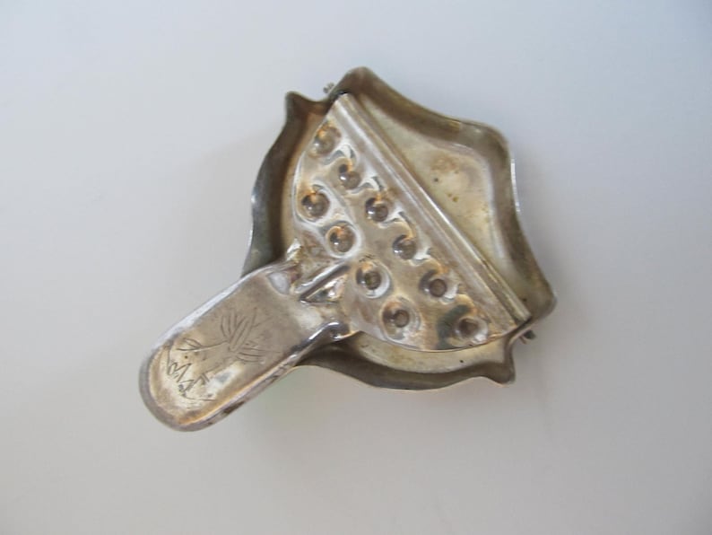 Op de afbeelding: Zilverkleurige metalen citruspers met een handvat en een getextureerd oppervlak. De citruspers heeft een reeks kleine gaatjes om citrusvruchten uit te persen. Het handvat heeft een klein boomontwerp en de tekst "M.A.K." erop gegraveerd.