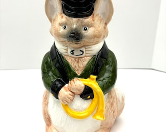 May include: A ceramic figurine of a fox wearing a green jacket, black hat, and white bow tie. The fox is holding a yellow horn in its paws.