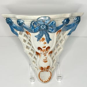 May include: White ceramic wall shelf with a decorative design of blue ribbons and orange floral accents. The shelf has a lattice pattern and a scalloped edge.