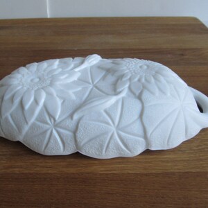 May include: White milk glass dish with a floral design featuring sunflowers and leaves. The dish has a scalloped edge and two handles.