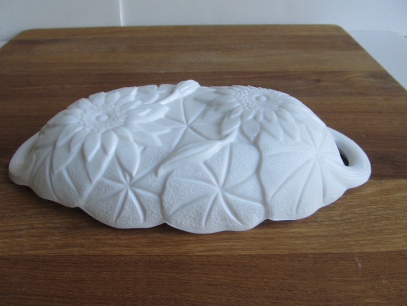 May include: White milk glass dish with a floral design featuring sunflowers and leaves. The dish has a scalloped edge and two handles.