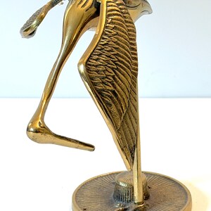 May include: A polished brass figurine of a bird with its head tucked downward, featuring detailed wing feathers and a long beak. The bird stands on a circular base with a radial sunburst pattern etched into the metal surface, creating a reflective finish.