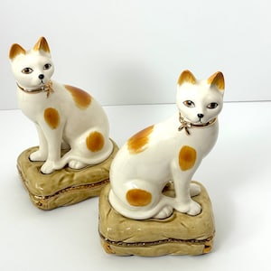 May include: Two white porcelain cat figurines with brown spots, each sitting on a gold-trimmed cushion. The cats have gold collars and black eyes.