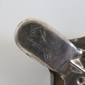 Op de afbeelding: Close-up van een zilverkleurig metalen voorwerp met een gegraveerd ontwerp. Het ontwerp toont een gestileerde vogel met een lange staart en een bloemmotief. Het voorwerp heeft een afgeronde vorm en een glad oppervlak.