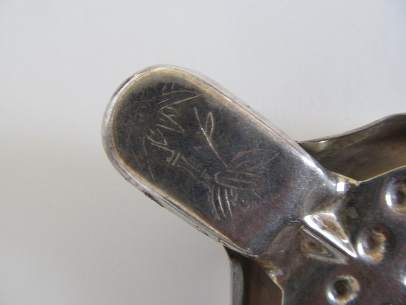 Op de afbeelding: Close-up van een zilverkleurig metalen voorwerp met een gegraveerd ontwerp. Het ontwerp toont een gestileerde vogel met een lange staart en een bloemmotief. Het voorwerp heeft een afgeronde vorm en een glad oppervlak.