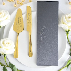 May include: A gold-colored cake server and knife set with engraved text and a decorative floral design. The set is displayed with a matching dark gray box. The text reads "Dave & Claire" and "24.05.2015".