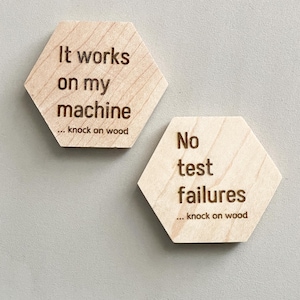 Puede incluir: Dos placas hexagonales de madera clara. Una dice "It works on my machine... knock on wood." La otra dice "No test failures... knock on wood." El texto es de color marrón oscuro.