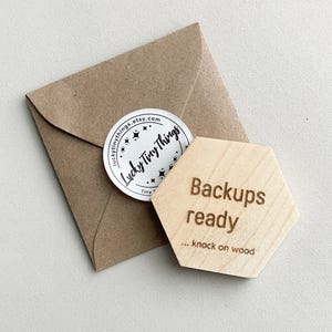 Peut inclure: Un hexagone en bois clair avec l'inscription "Backups ready ... knock on wood". Il est posé à côté d'une enveloppe marron avec un autocollant rond "Lucky Tiny Things".