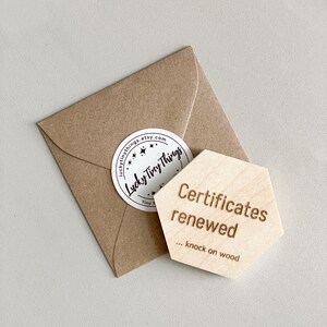 May include: A small, brown paper envelope with a white sticker that reads "luckytinythings.etsy.com". Next to it is a wooden hexagon with the words "Certificates renewed... knock on wood" engraved on it.