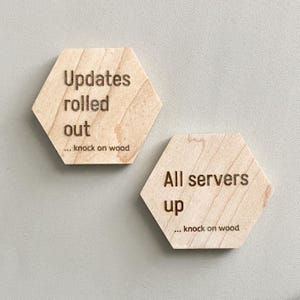 Könnte beinhalten: Zwei hellfarbene, sechseckige Untersetzer aus Holz. Einer der Untersetzer trägt die Aufschrift "Updates rolled out... knock on wood". Der andere lautet "All servers up... knock on wood". Die Untersetzer liegen auf einer hellgrauen Oberfläche.