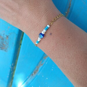 Peut inclure: Un bracelet à chaîne dorée orné d'une perle centrale bleu foncé en forme d'œil protecteur, entourée de perles blanches et turquoise alternées. La fine chaîne en métal est reliée à la section perlée, formant un accessoire simple et équilibré.