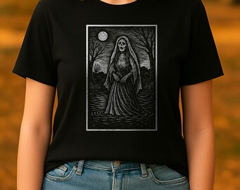 May include: Black t-shirt featuring a detailed black and white illustration of a spectral figure in a flowing gown, standing in water under a full moon. The artwork is framed with a silver border, creating a gothic or Halloween theme.