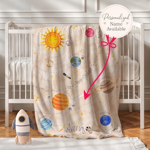 May include: A beige baby blanket featuring a colorful solar system design with planets, stars, and the sun. The blanket is draped over a white crib. The name "Avery" is printed on the blanket. A plush rocket toy sits on the floor.