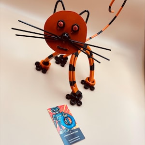 May include: A whimsical metal cat sculpture in orange and black. The cat has a round head, long whiskers, and a striped tail. The eyes are red. A business card with a robot graphic is in the foreground.