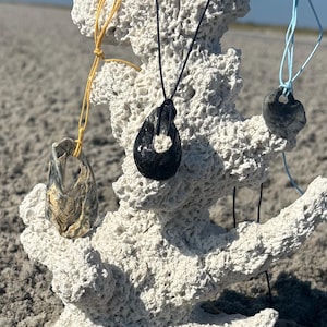 以下が含まれることがあります： 白いサンゴの構造に展示された、天然石のペンダントが付いた3つの手作りのペンダントネックレス。ネックレスは黒、黄、青のコードが付いています。さまざまな貝殻がサンゴの根元に配置されています。
