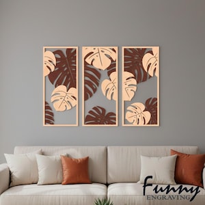 Könnte beinhalten: Dreiteilige Wandkunst mit einem botanischen Design. Jedes Paneel hat einen hellbraunen Rahmen und einen grauen Hintergrund. Das Design umfasst hellbraune und dunkelbraune Monstera-Blätter. Der Text "Funny Engraving" befindet sich unten rechts.