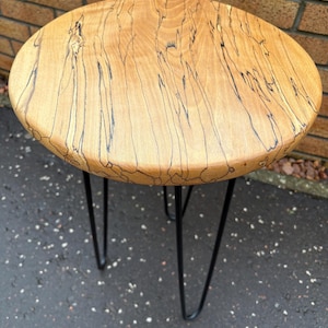 May include: A round wooden side table with a light brown top featuring dark, linear grain patterns. The table is supported by three black hairpin legs, creating a mid-century modern aesthetic. The table's top is approximately 18 inches in diameter.