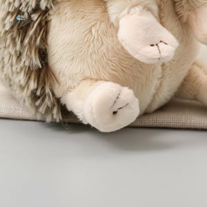 May include: A soft toy hedgehog with beige fur and brown quills. The toy features small, cream-coloured paws with stitched details. The hedgehog is resting on a neutral-coloured surface, creating a cosy and inviting appearance.