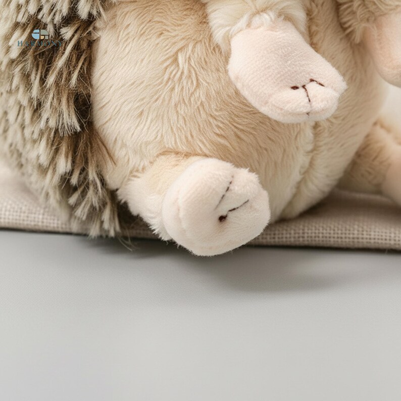 May include: A soft toy hedgehog with beige fur and brown quills. The toy features small, cream-coloured paws with stitched details. The hedgehog is resting on a neutral-coloured surface, creating a cosy and inviting appearance.