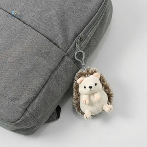 May include: A grey backpack with a silver zip and a small, plush hedgehog keyring. The keyring is cream-coloured with brown quills and small, black eyes. The backpack is made of a textured fabric.