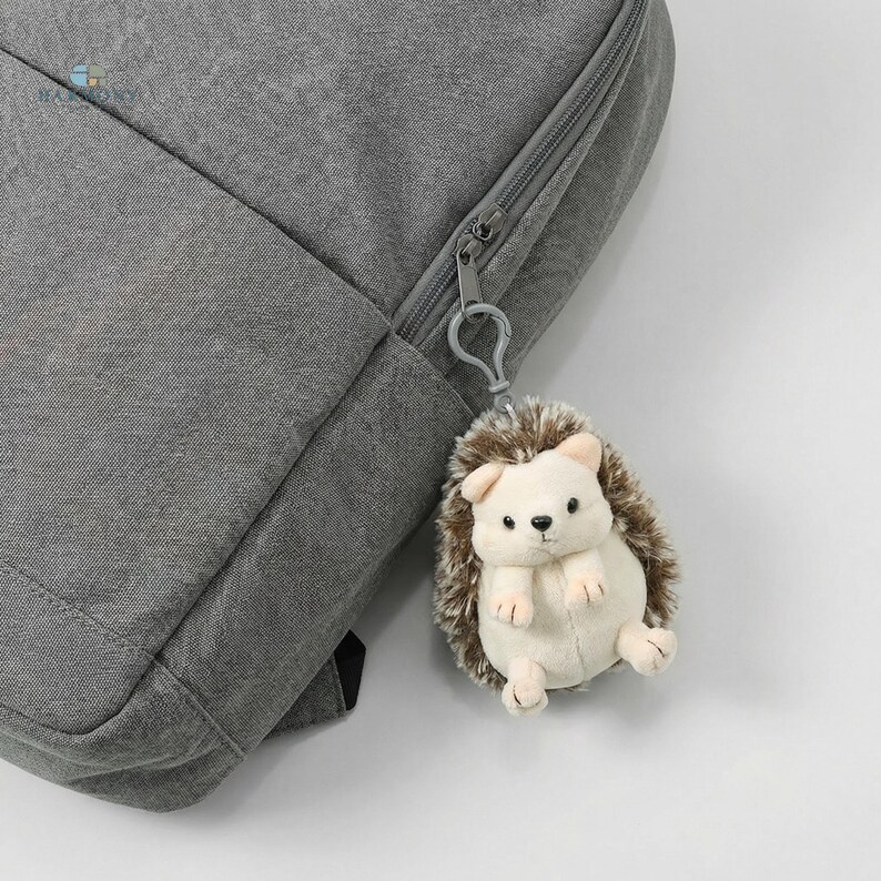 May include: A grey backpack with a silver zip and a small, plush hedgehog keyring. The keyring is cream-coloured with brown quills and small, black eyes. The backpack is made of a textured fabric.