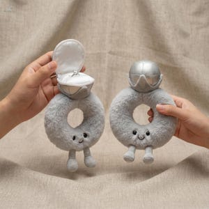 May include: Two plush gray ring toys, one with an open ring box holding a diamond ring. The other ring toy has a silver top. Both have smiling faces and small legs. The toys are designed to look like engagement rings.
