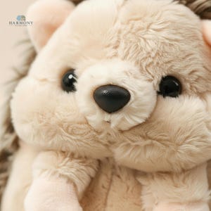 May include: A close-up shot of a soft, plush hedgehog toy. The toy has beige fur and darker brown spines. It has large, black eyes and a black nose. The toy is designed to be cuddly and is suitable for children.