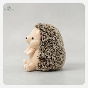 May include: A soft toy hedgehog, shown seated. It features a light beige face, paws, and belly, with a dark brown and beige spiky back. The hedgehog has small black eyes and a pink nose. The Harmony logo is visible in the top left corner.