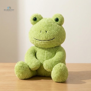 Könnte beinhalten: Ein grünes Plüschfrosch-Spielzeug mit großen, runden Augen und einem lächelnden Mund. Der Frosch besteht aus einem weichen, strukturierten Material und sitzt auf einer Holzoberfläche. Das Spielzeug hat ein freundliches, cartoonartiges Aussehen.