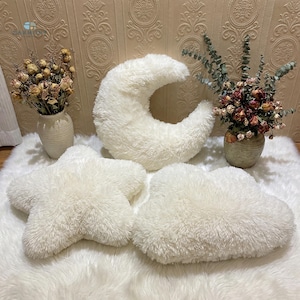 Puede incluir: Un conjunto de cojines decorativos blancos y esponjosos con forma de estrella, media luna y nube. Los cojines están colocados sobre una superficie blanca texturizada, con jarrones de flores secas en el fondo.