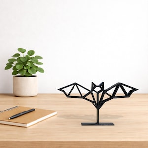 Peut inclure: Une sculpture de chauve-souris géométrique noire avec un design triangulaire, sur une petite base noire. La chauve-souris est exposée sur une surface en bois, avec un carnet, un stylo et une plante en pot en arrière-plan.