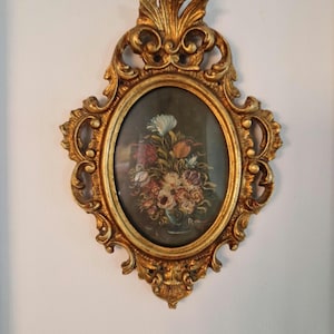 Può includere: Un'opera d'arte da parete di forma ovale con un dipinto di natura morta floreale racchiuso in una cornice dorata ornata con intricati motivi a volute e dettagli a forma di foglia nella parte superiore e inferiore. Il dipinto raffigura un bouquet.