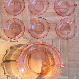 Könnte beinhalten: Ein Set aus sechs rosa Dessertschalen aus Glas und einer Servierschüssel. Die Schalen haben einen gewellten Rand und kleine Griffe. Das Glas ist leicht transparent, und die Schalen sind auf einer Holzoberfläche angeordnet.