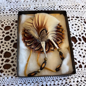 Puede incluir: Broche decorativo de plumas en una caja negra. El broche presenta un abanico de plumas crema y marrón con un acento de metal plateado. Tiene un colgante de cuentas con cuentas negras y marrones y un cordón beige.