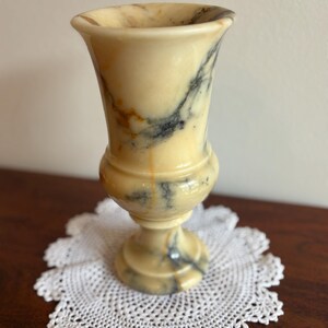 Può includere: Un vaso decorativo in marmo color crema con venature grigio scuro e marroni. Il vaso ha una parte superiore svasata, una sezione centrale arrotondata e una base a piedistallo. È appoggiato su un centrino bianco all'uncinetto. Il vaso è alto circa 15 cm.