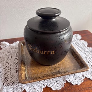 Op de afbeelding: Een zwarte keramische tabakspot met een deksel, met het woord "Tobacco" in gouden letters. De pot staat op een rechthoekige, gespikkelde tray. De achtergrond omvat een wit kanten kleedje en een houten oppervlak.