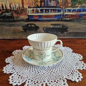 Peut inclure: Une tasse et une soucoupe blanches avec des motifs floraux reposent sur un napperon blanc en crochet. La tasse a un bord cannelé et une anse délicate. La soucoupe et la tasse présentent un motif floral avec des accents roses et bleus. En arrière-plan, une peinture d'une scène de ville.