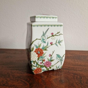 Könnte beinhalten: Eine weiße Porzellankanne mit quadratischem Deckel, verziert mit farbenfrohen Blumen- und Vogelmotiven. Die Kanne zeigt ein Design aus roten, rosa und weißen Blumen, grünen Blättern und blauen Vögeln, die auf Ästen sitzen. Der Deckel hat einen grün-weißen Rand.