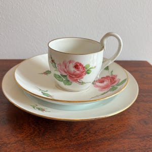 Op de afbeelding: Een vintage theeservies bestaande uit een witte kop en schotel en een bord. Elk stuk heeft een roze rozenmotief en gouden randen. De kop heeft een gebogen oor. De set staat op een houten oppervlak.